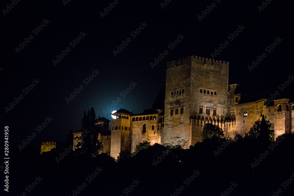 A view of the Alhambra with the moon, a Moorish castle, fortress and palace, lit up at night in Granada, Spain