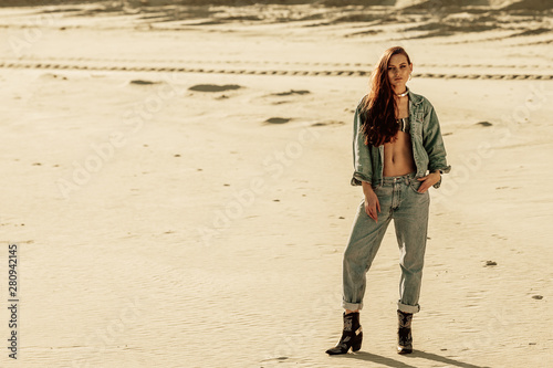 Outdoor full-length portrait of young fashionable woman with long hair,  wearing total denim outfit: jacket, jeans, trendy cowboy boots, posing in the desert, at sunset. Copy, empty space for text