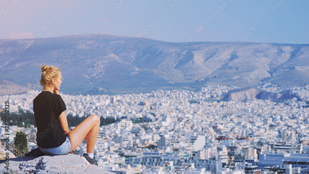 Young tourist woman sitting on top of mountain and looking at a beautiful landscape cityscape Athens Greece. Adult girl tourist relax on hill overlooking Athens in summer. Famous Athens city in Europe