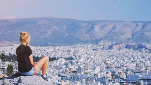 Fototapeta Naklejka Na Ścianę i Meble -  Young tourist woman sitting on top of mountain and looking at a beautiful landscape cityscape Athens Greece. Adult girl tourist relax on hill overlooking Athens in summer. Famous Athens city in Europe
