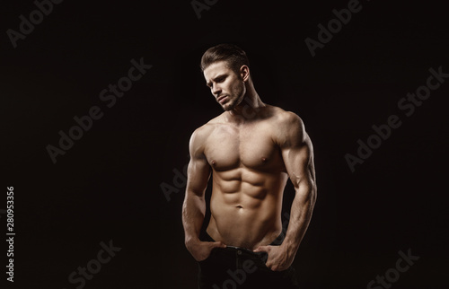 Muscular model sports young man on dark background. Fashion portrait of strong brutal guy with a modern trendy hairstyle. Sexy torso. Male flexing his muscles.