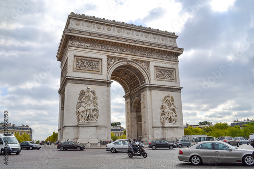 Front view of Arch of Triumph in Paris - France, with a cloudy sky and traffic of cars, bikes and the city behind it.