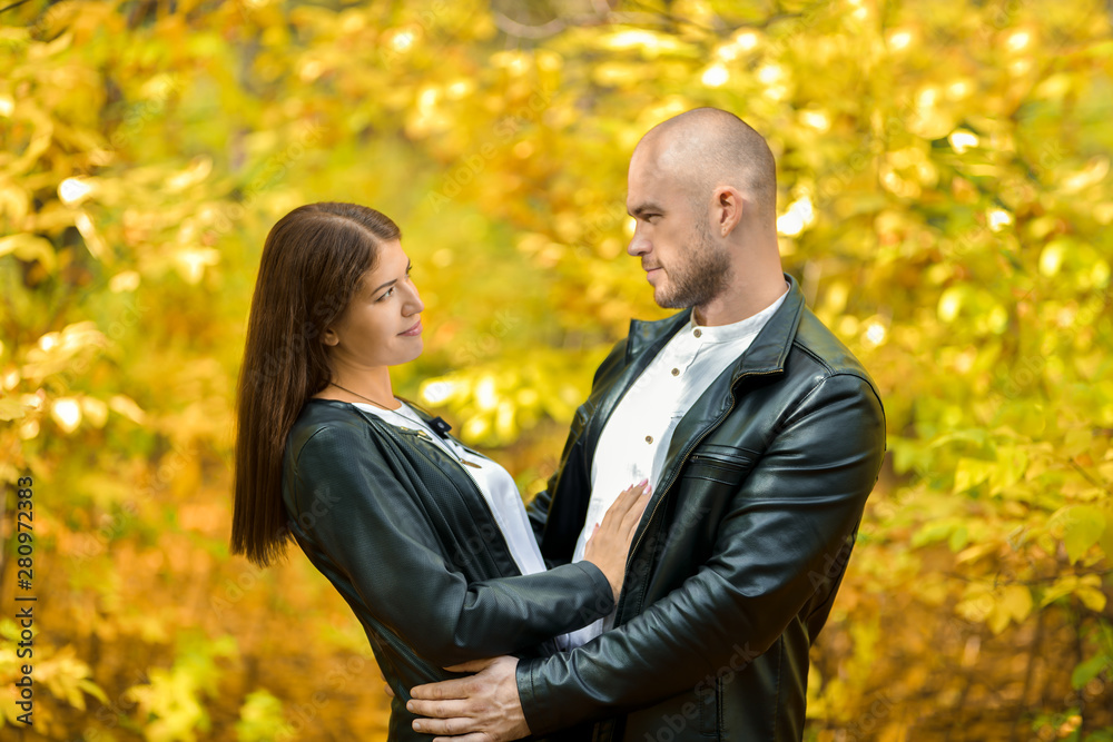 Autumn forest. A dark-haired beautiful girl and a bald brutal man in leather jackets tenderly look at each other. A man and a woman hugging. Yellow autumn background. Horizontal photography Autumn forest. A dark-haired beautiful girl and a bald brutal man in leather jackets tenderly look at each other. A man and a woman hugging. Yellow autumn background. Horizontal photography