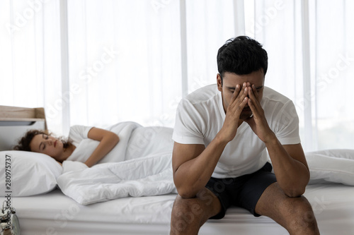 Upset depressed young man sitting on the edge of bed against his wife lying on the bed. Relationship difficulties.