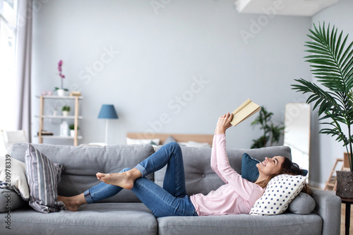 Beautiful smiling woman reading a book and lying on the sofa in the living room.