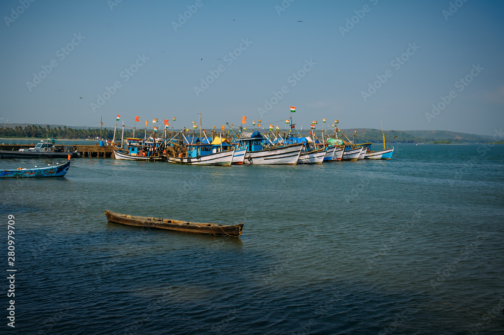 Colorful fishing boats with blue and white hulls and Indian flags on the masts, on the fishing pier in Goa. Wooden boats in the light of morning sun, birds flying in the sky. India independence day.