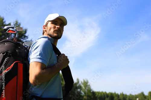 Portrait of male golfer carrying golf bag while walking by green grass of golf club.