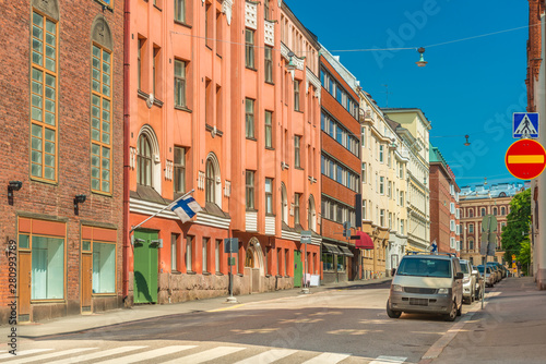 View of an empty street in Helsinki, Finland. Colored historical buildings with Finnish flags on the facades, parked cars and clear blue sky