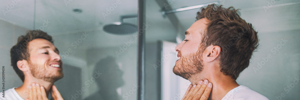 Fototapeta premium Man putting after shave perfume lotion or skin care cream for sensitive skin after shaving beard looking in bathroom mirror on neck. Male beauty, beard care skincare concept.