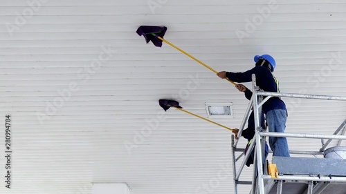 Low angle view of 2 workers on scaffolding using mop sticks to cleaning white ceiling roof of petrol station
