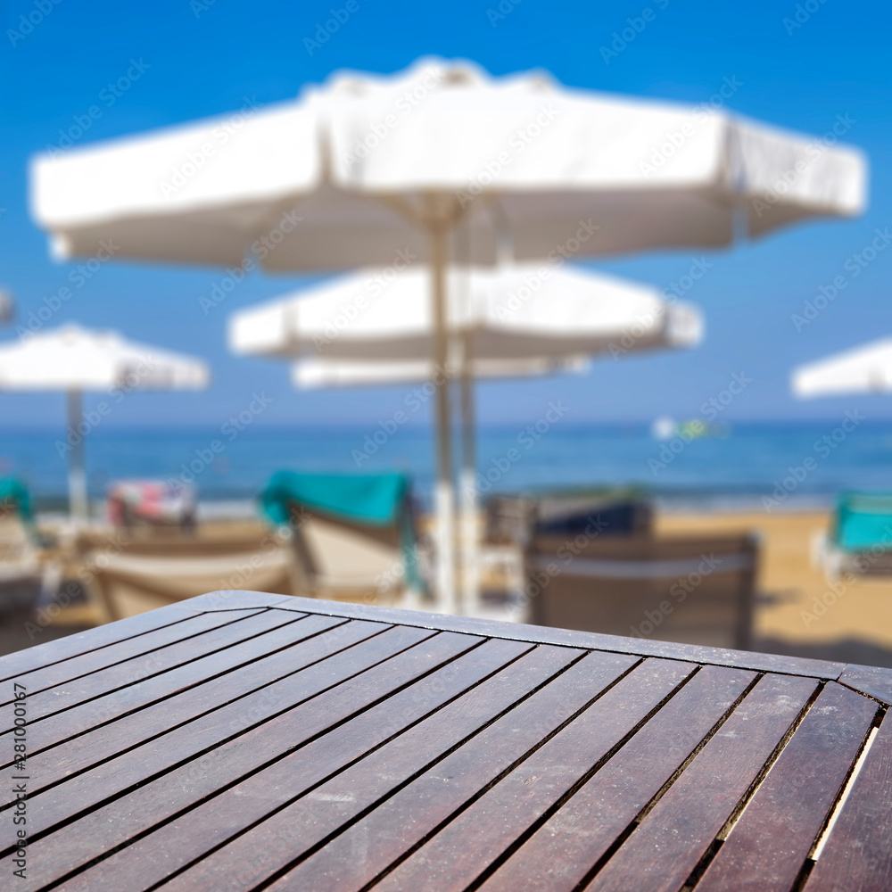 Table background on a sandy beach view with blurred sun beds and umbrellas in distance.
