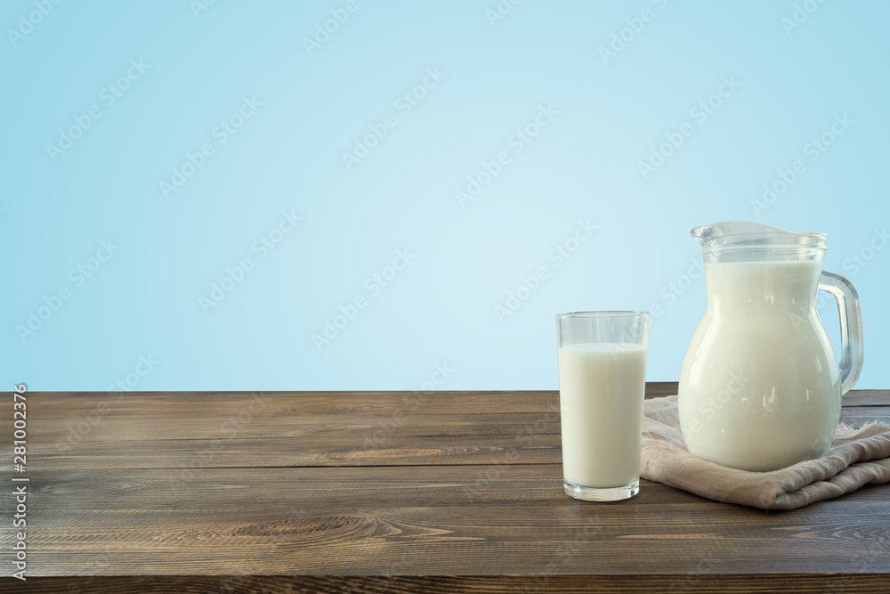 Glass of fresh milk and jug on wooden tabletop with blue wall as background.