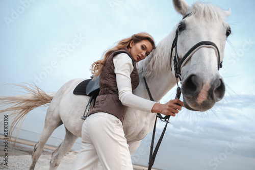 Attractive and beautiful young woman wearing stylish jockey outfit is holding the reins and posing with the white horse on the coast near the water. An attractive rider is posing outdoors. Сountryside