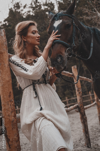 Beautiful and tender young woman wearing the dress is embracing and stroking the horse on the ranch. An attractive rider is posing outdoors near the saddle. Summertime, nature landscape, countryside