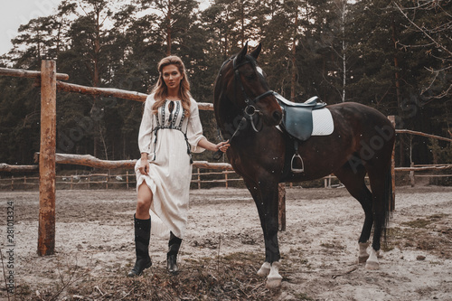 Pretty and sensual young woman wearing the dress is holding the reins and posing with the horse on the ranch near the saddle. An attractive rider is posing outdoors. Forest, nature landscape