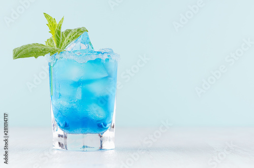 Tropical fresh cocktail with blue curacao liqueur, ice cube, sugar rim, green mint in frozen shot glass on soft light white, pastel mint color background.