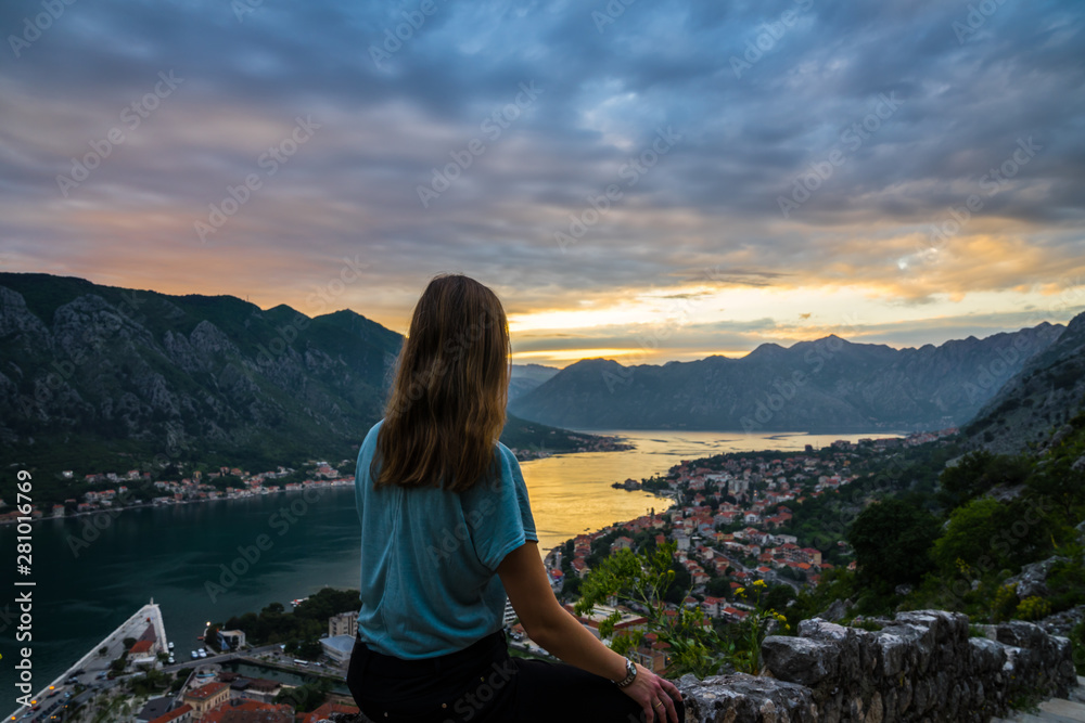 Montenegro, Young blonde beauty woman sitting on stone wall above kotor bay city in beautiful orange sunset twilight dawning atmosphere in summer vacation season