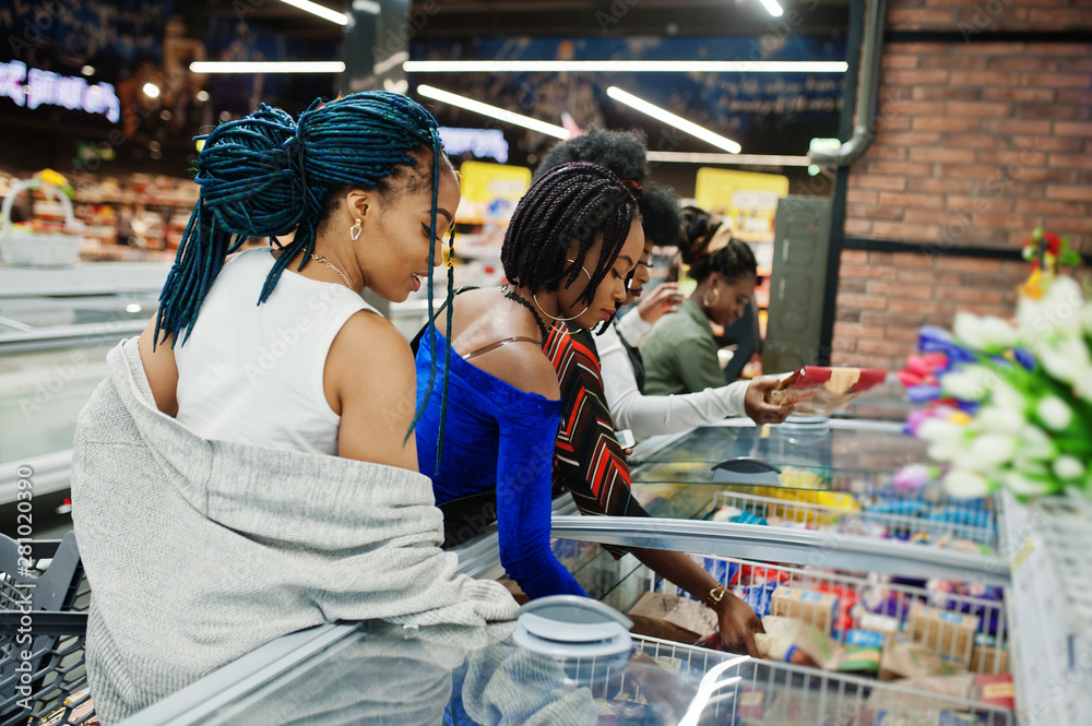 Obraz premium Group of african womans with shopping carts near refrigerator choose packs of dumplings in the supermarket.