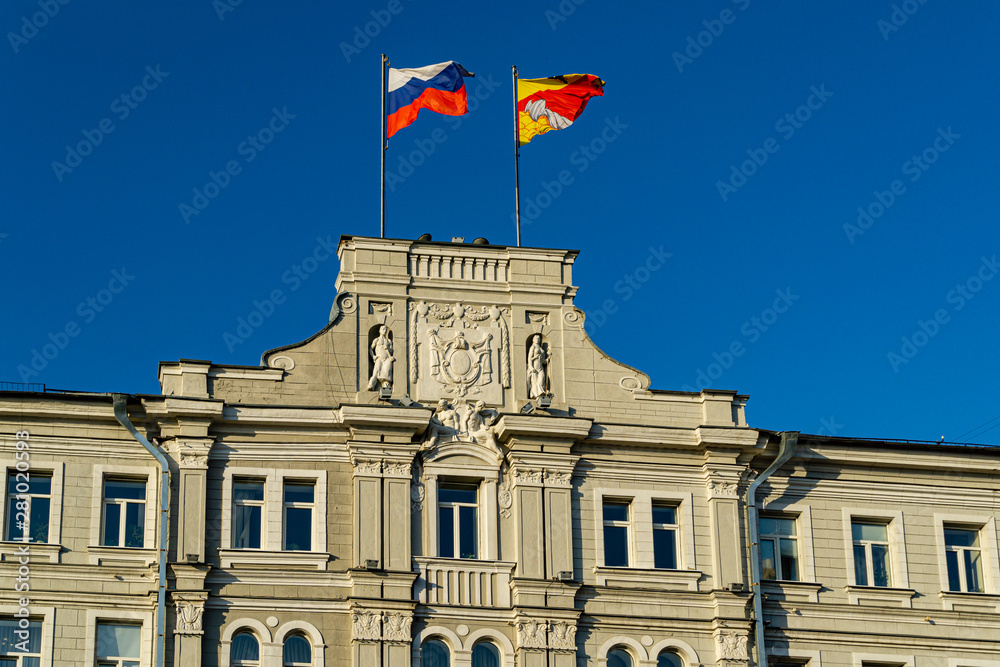 Close-up of top decoration of main facade Voronezh city administration building on Plekhanovskaya Street 10. Building was built in 1914-1915. Flags of Russia and region flying against blue sky