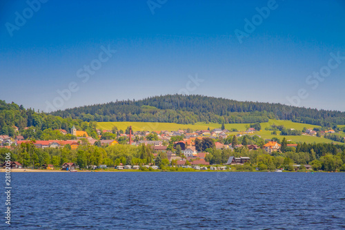 The Lipno Dam - Horni Plana, Czech Republic, in a bright summer day. The lake is calm, has clean blue water. There are no clouds in the sky. Lot of trees on the background.