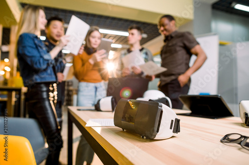 Blurred image of five multiethnical business persons are developing a project using modern gadgets and virtual reality goggles. Focus on the VR goggles on the table. technologies of the future