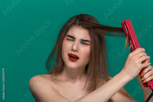 Portrait of beautiful young woman with bright color make-up having probems doing hair style herself with a purple straightening iron photo