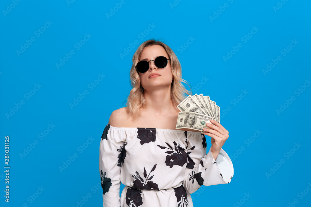 Portrait of a girl with curly blond hair in a white dress and dark sunglasses standing on a blue background. Happy model fans herself with a bundle of dollars with dreamy look.