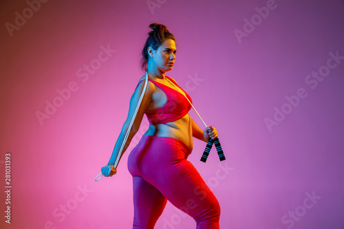 Young caucasian plus size female model's training on gradient purple background in neon light. Doing workout exercises with the jump rope. Concept of sport, healthy lifestyle, body positive, equality.