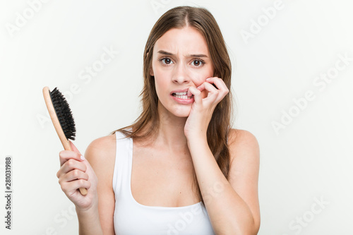 Young caucasian woman holding a hair brush biting fingernails, nervous and very anxious.