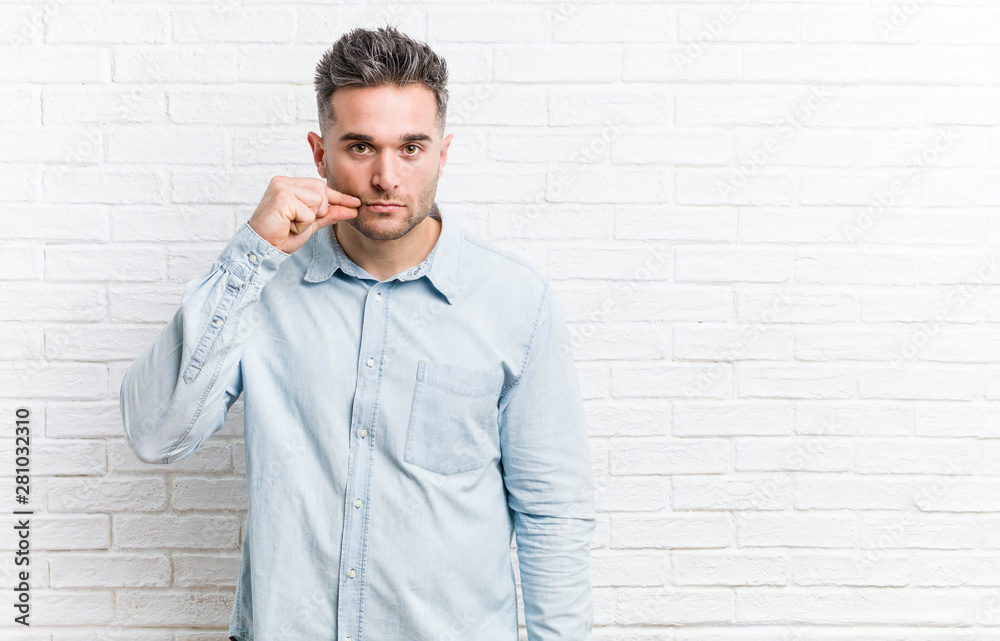 Young handsome man against a bricks wall with fingers on lips keeping a secret.