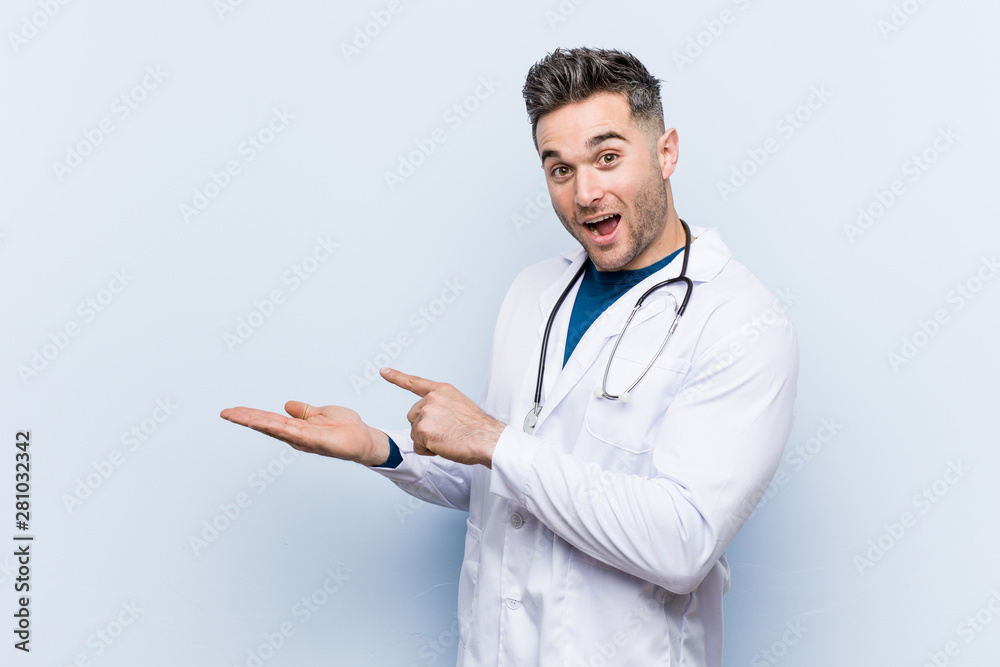 Young handsome doctor man excited holding a copy space on palm. Young handsome doctor man excited holding a copy space on palm.