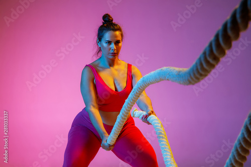 Young caucasian plus size female model's doing exercises on gradient purple background in neon light. Cross training with the rope. Concept of sport, healthy lifestyle, body positive, equality. photo