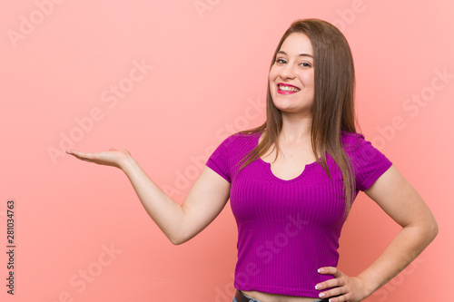 Young hispanic woman against a pink wall showing a copy space on a palm and holding another hand on waist.