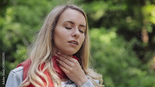 Sick young woman wearing red scarf feeling bad suffering from throat pain, sore throat, standing in the park. Painful Swallowing, health concept.