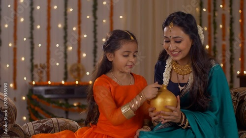 Beautiful mother teaching her cute daughter the concept of money saving - Parenting and Financial . South Indian daughter learning how to save money from her mother  putting a coin in a gullak on D... photo