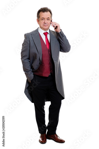 Full body portrait of handsome confident caucasian mature businessman 55-60 years old in grey tuxedo and red vest. Rich senior man talk by phone and looking to camera on isolated white background