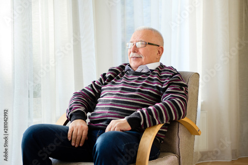 Portrait of smiling and confident senior man 70-75 years old with eyeglasses relaxing in armchair near light window at home. Elder male looking at window