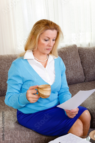 Business middle-age woman 60-65 years old sit on grey sofa and works at home. Senior female reads the letter with a surprised expression with cup in hand.