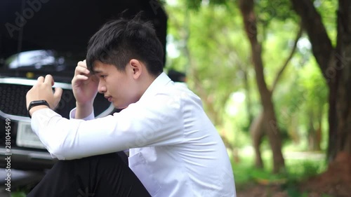 A young Asian man sitting in front of a broken car at the side of the road