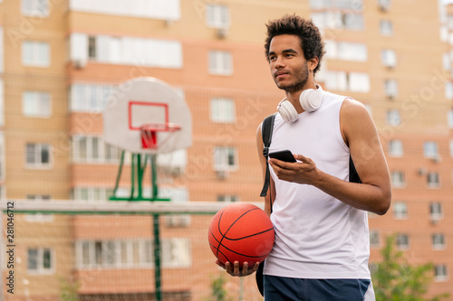 Young mixed-race athlete with ball and smartphone texting after game © pressmaster