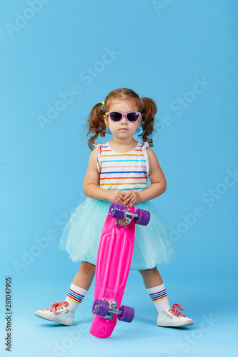 Cool little toddler girl in bright clothes and sunglasses carrying a skateboard over his shoulder and looking at the camera isolated on blue background