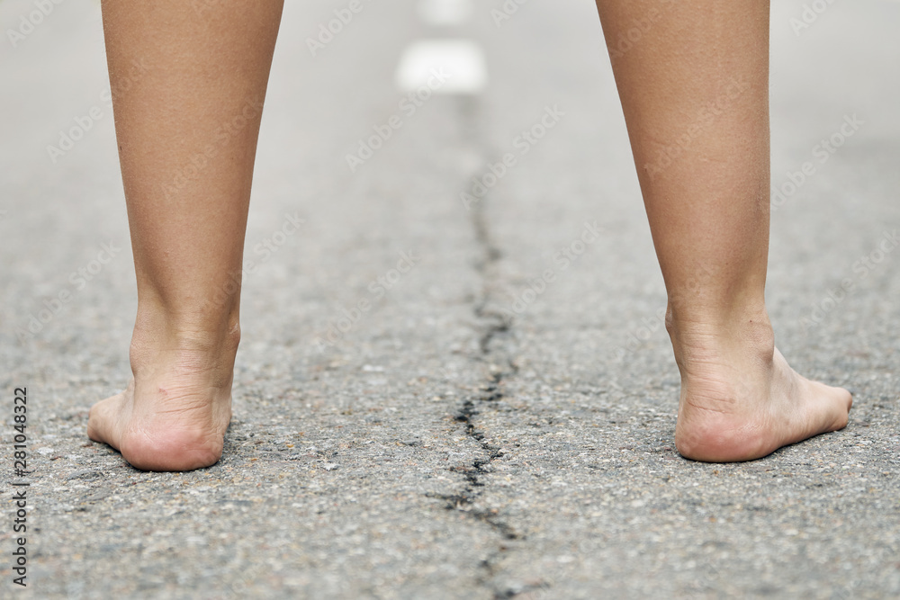 Rear view of bare feet of a young girl standing on the asphalt road close up. Dividing road lines are visible far away between legs.