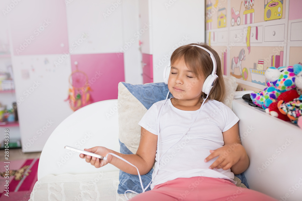 Close up portrait of little caucasian adorable girl listening to music in earphones in cozy bedroom smiling and relaxing. Happiness, leisure, children lifestyle, childhood, gadget,technology concept Close up portrait of little caucasian adorable girl listening to music in earphones in cozy bedroom smiling and relaxing. Happiness, leisure, children lifestyle, childhood, gadget,technology concept
