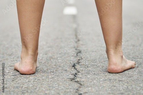 Rear view of bare feet of a young girl standing on the asphalt road close up. Dividing road lines are visible far away between legs.