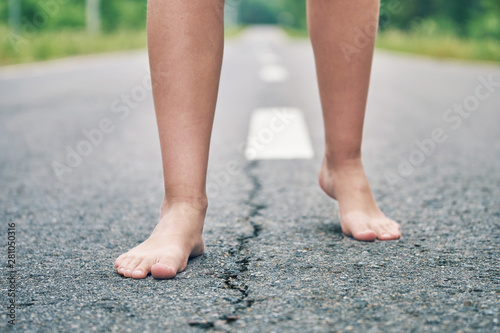 Front view of bare feet of a young girl walking along the asphalt road close up. Dividing road lines are visible far away between legs.
