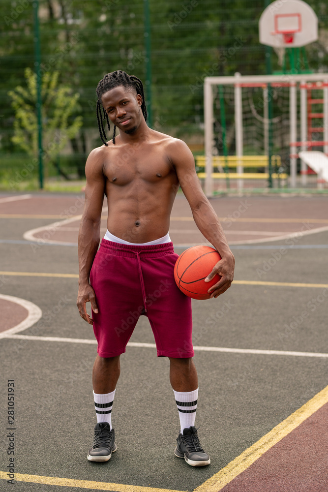 Shirtless African muscular guy with ball standing on basketball court