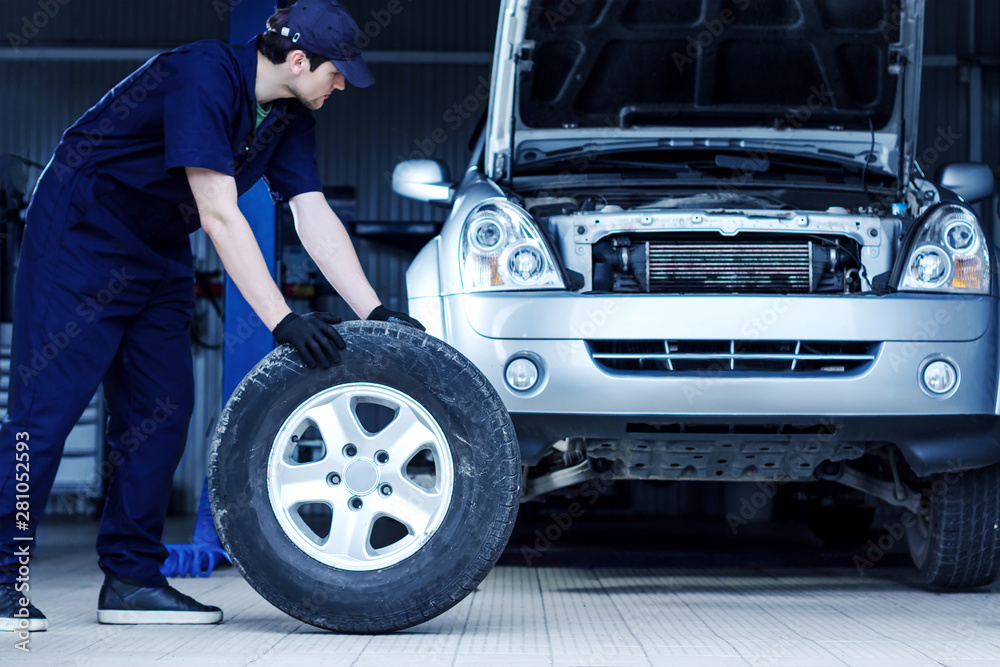 Mechanic in blue jumpsuit is repairing car at modern service station. Repairman is rolling wheel on floor of workshop auto repair shop. Silver vehicle on background. Tire fitting concept. Mechanic in blue jumpsuit is repairing car at modern service station. Repairman is rolling wheel on floor of workshop auto repair shop. Silver vehicle on background. Tire fitting concept.