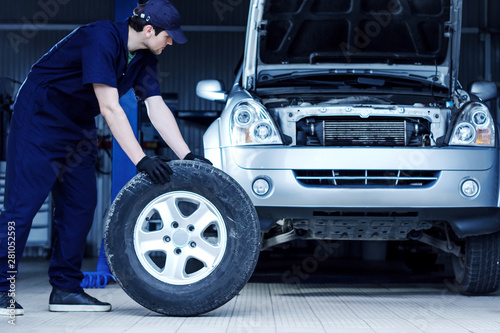 Mechanic in blue jumpsuit is repairing car at modern service station. Repairman is rolling wheel on floor of workshop auto repair shop. Silver vehicle on background. Tire fitting concept.