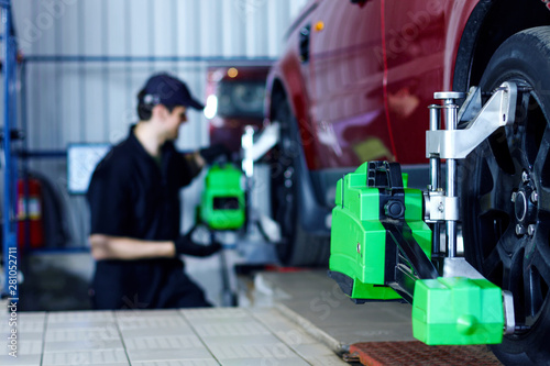 Man mechanic is repairing red car at modern service station. Repairer in blue jumpsuit and cap is adjusting sensors on wheels. Process of wheels alignment camber check in workshop auto repair shop.