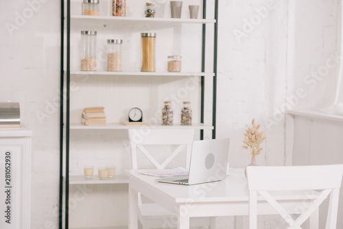 light kitchen with laptop on white table, chairs and rack with jars